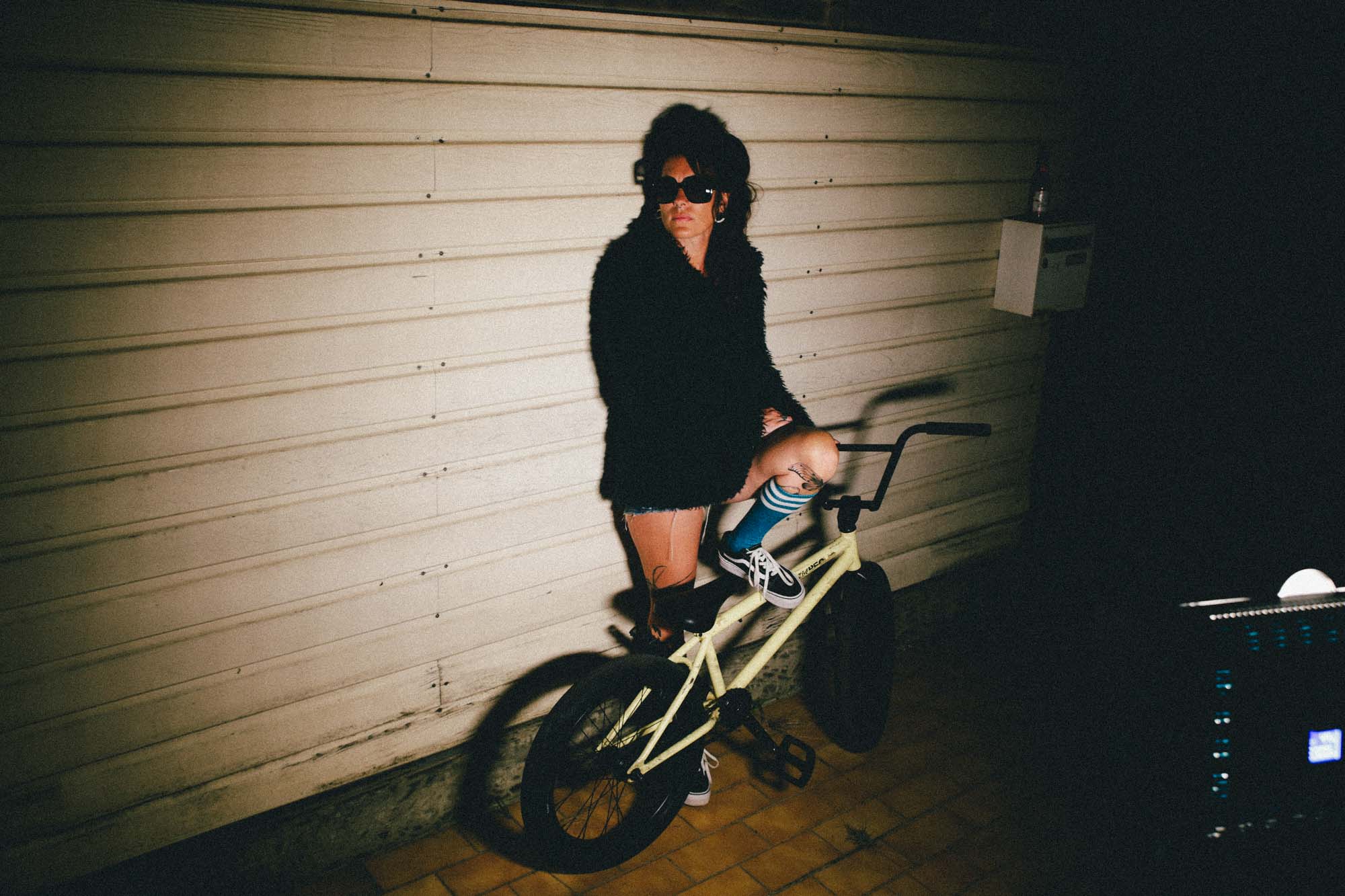 une juene femme , le regard de côté avec des lunettes noires, jambe posée sur un BMX, avec un air froid. photographie couleur, ton jaune