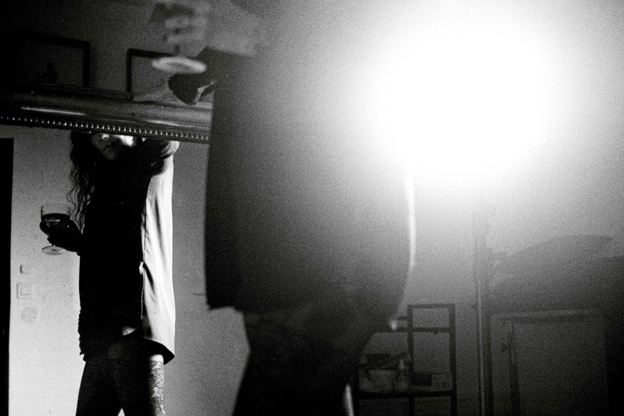 une femme tient un verre à la main et se regarde dans le mirroir aors qu'un lumière forte illumine la scéne, photo argentique noir et blanc