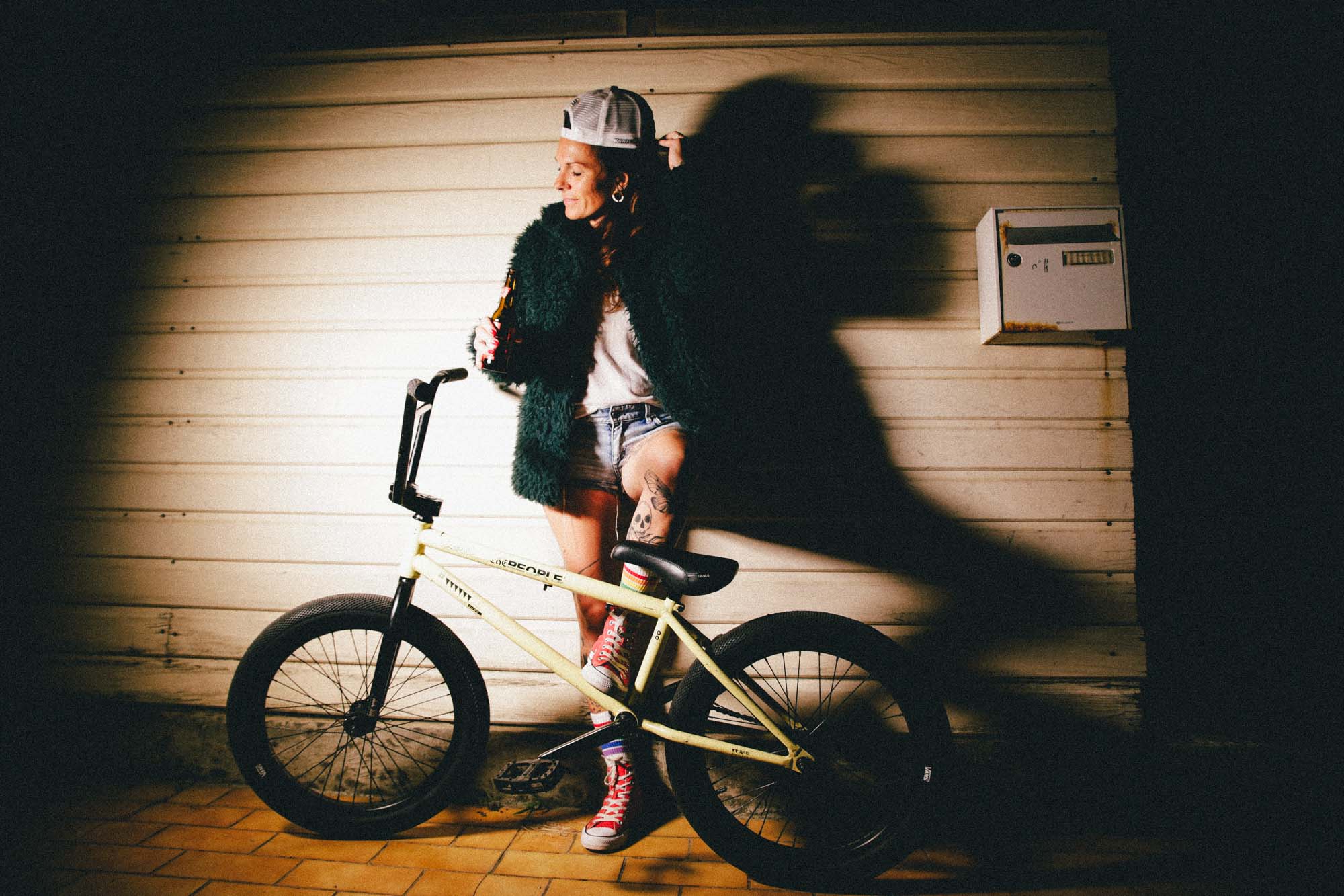 photographie couleur qui représente une femme adossée à un mur rouillé, qjiu pose ses pieds sur un BMX, elle remet sa casquette en place, une lumière artificielle jaune de côté apporte des ombres à la scène