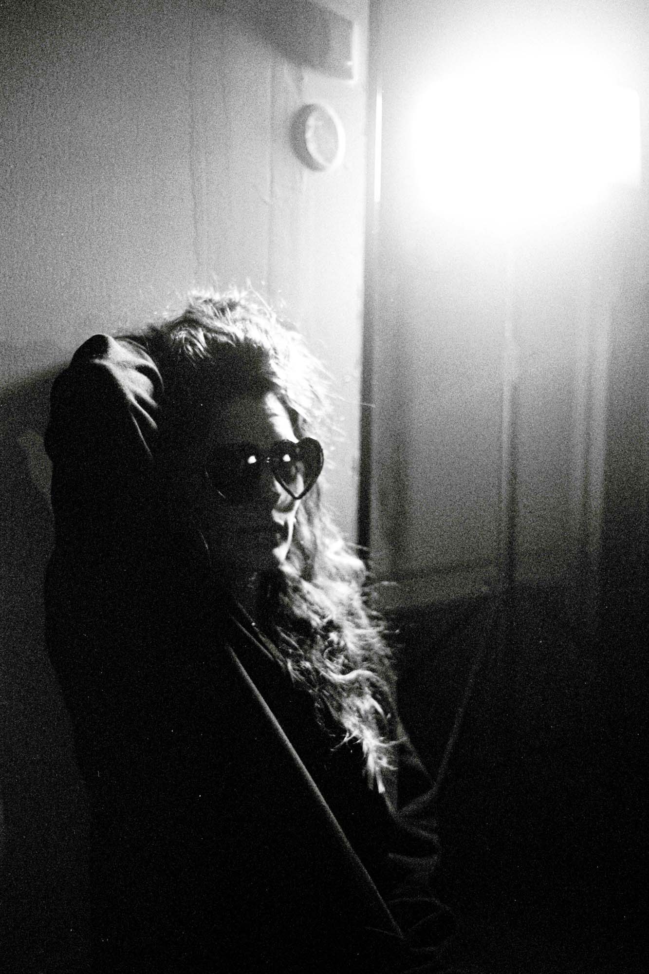 une femme se tient les cheveux , elle est adossée au mur avce des lunettes en forme de coeur, une lumière artificielle l'illumine, photographie argentique en noir et blanc
