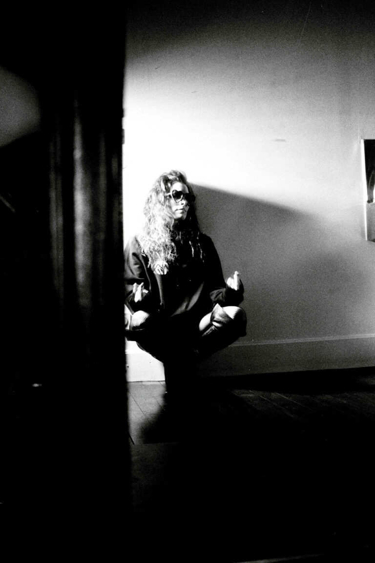 femme assisie qui me fait un signe avec les doigts , elleporte des lunnettes et est adossée au mur, photographie argentique noir et blanc