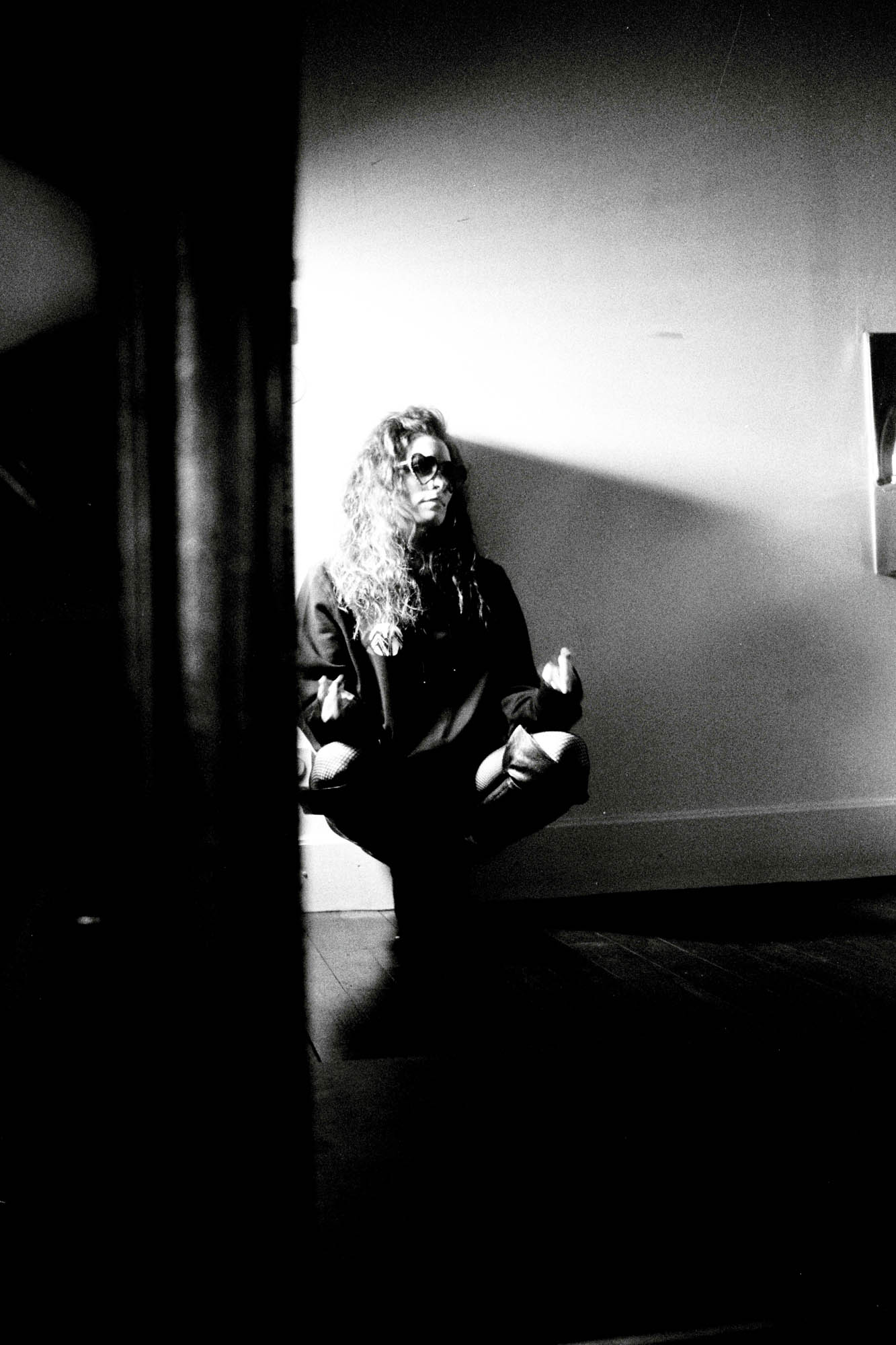 femme assisie qui me fait un signe avec les doigts , elleporte des lunnettes et est adossée au mur, photographie argentique noir et blanc