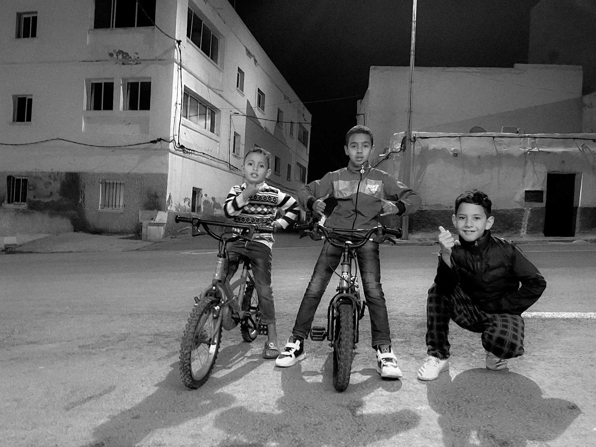 trois enfants posent devant leurs vélos un soir, on distingue les habitations derrière, ils sont fiers de leurs BMX, photographie noir et blanc