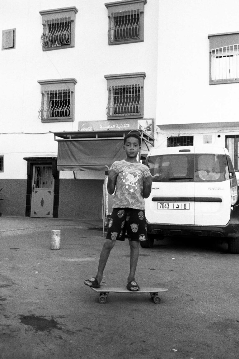 un enfant au maroc fait un signe de doigts, il est en tongue et monte sur un skateboard en bas d'un immeuble