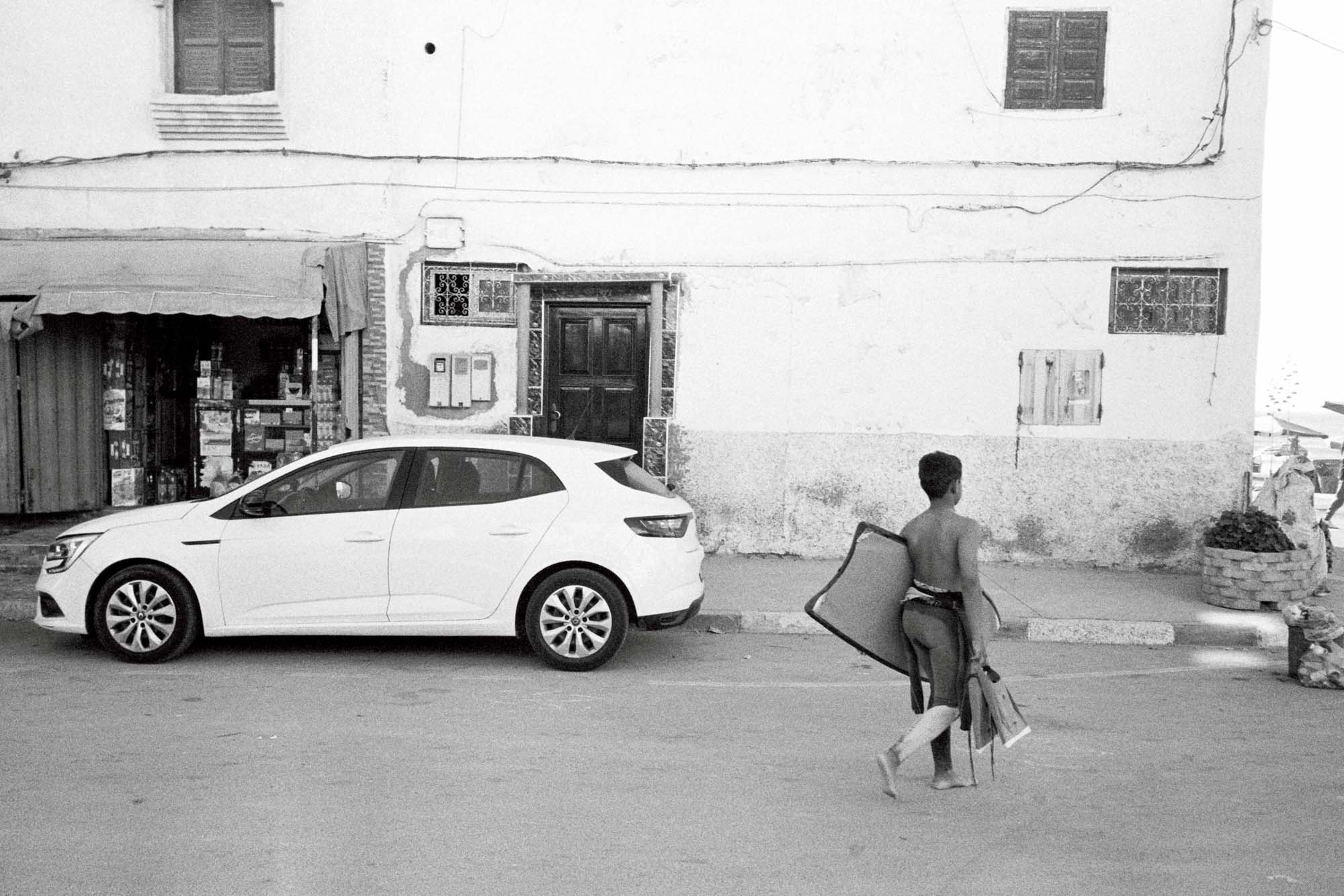 vue de dos d'un jeune enfant qui marche avec son bodyboard et ses palmes devant une façade marocaine et une boutique un peu délabrée en noir et blanc, argentique