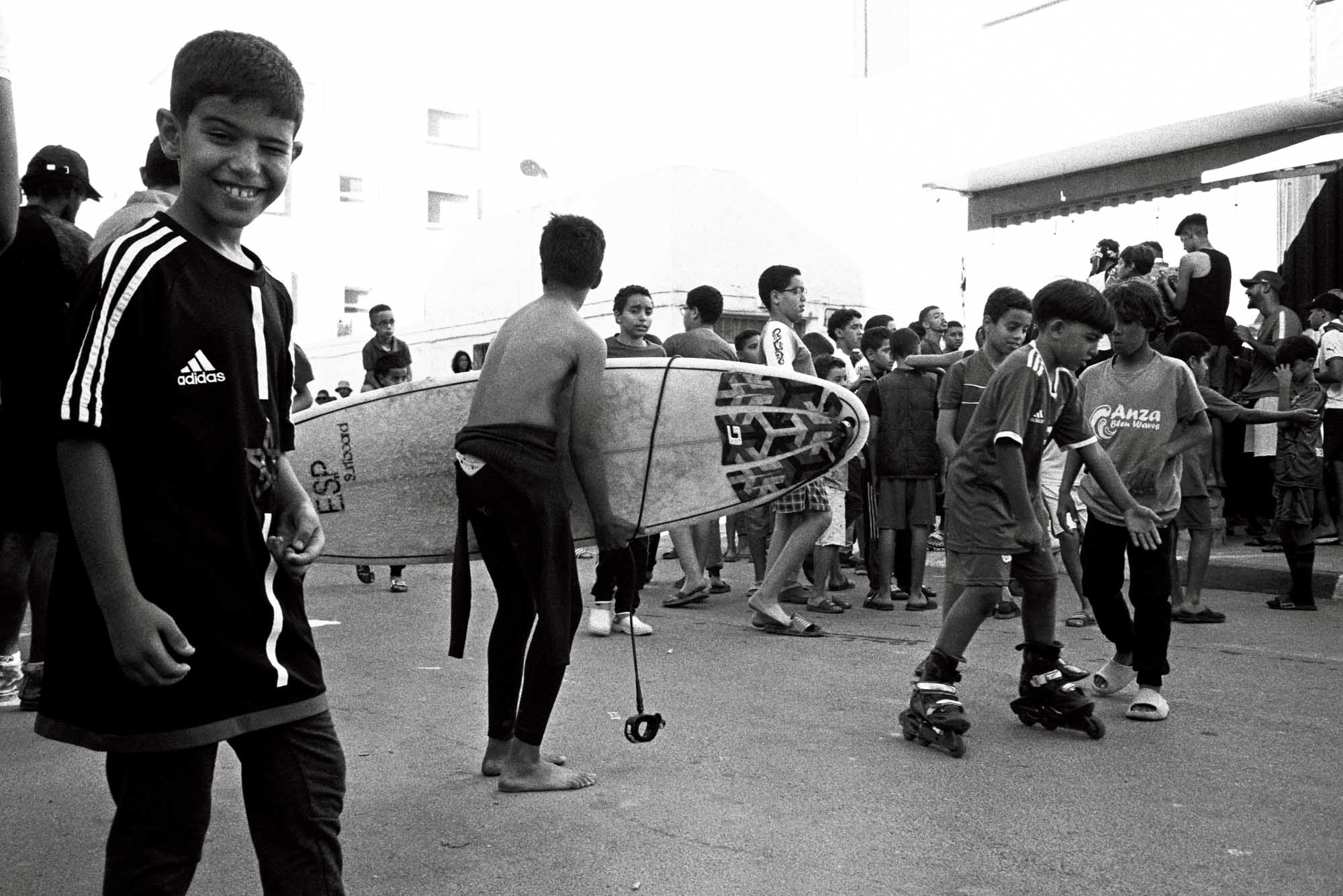 une foule de juenes enfants marocains sont dans la rue, on voit un qui porte son surf, un autre en roller et un dernier qui me regarde avec le sourire et une foule derrière