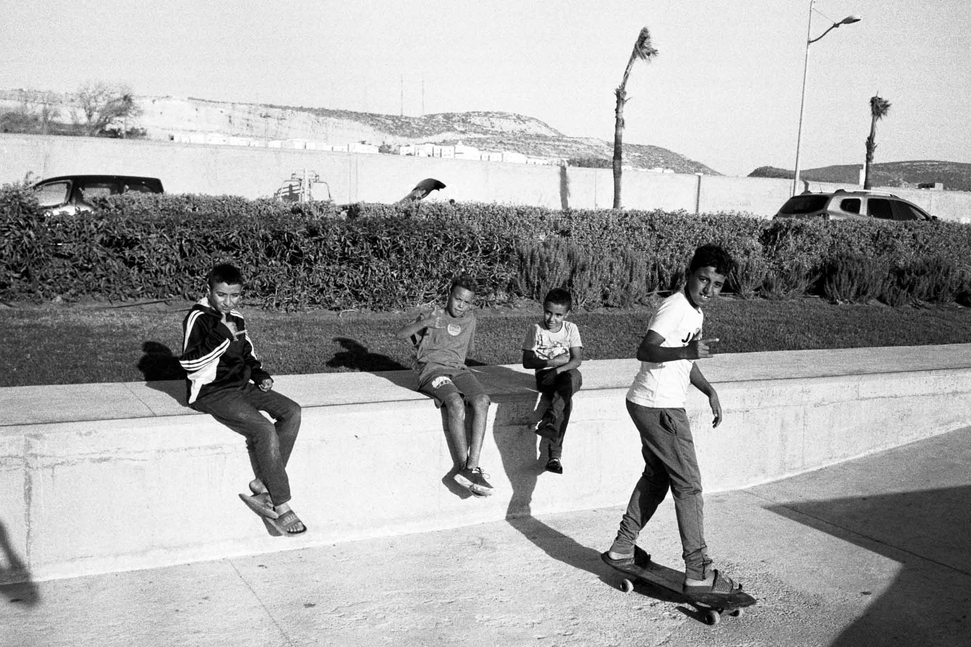 un groupe de jeunes marocains assis au skatepark dont le plus grand sur un skateboard en tongue qui me fait un signe de la main et me regarde, les aurte sont assis sur un muret derrière