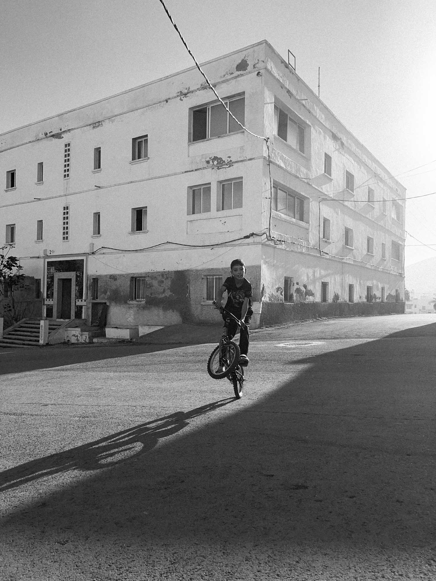un enfant au maroc fait une roue arrière avce son vélo au coucher de soleil, on distingue son ombre et celle du vélo, en noir et blanc