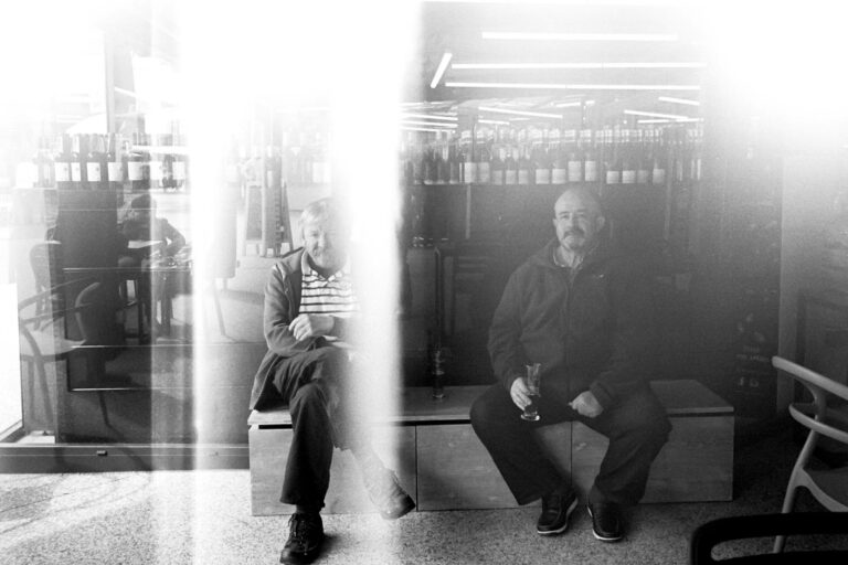 photographie noir et blanc argentique de deux vieux assis devant un magasin et qui boivent une bière. La photo a une bande blanche due au début de rouleau