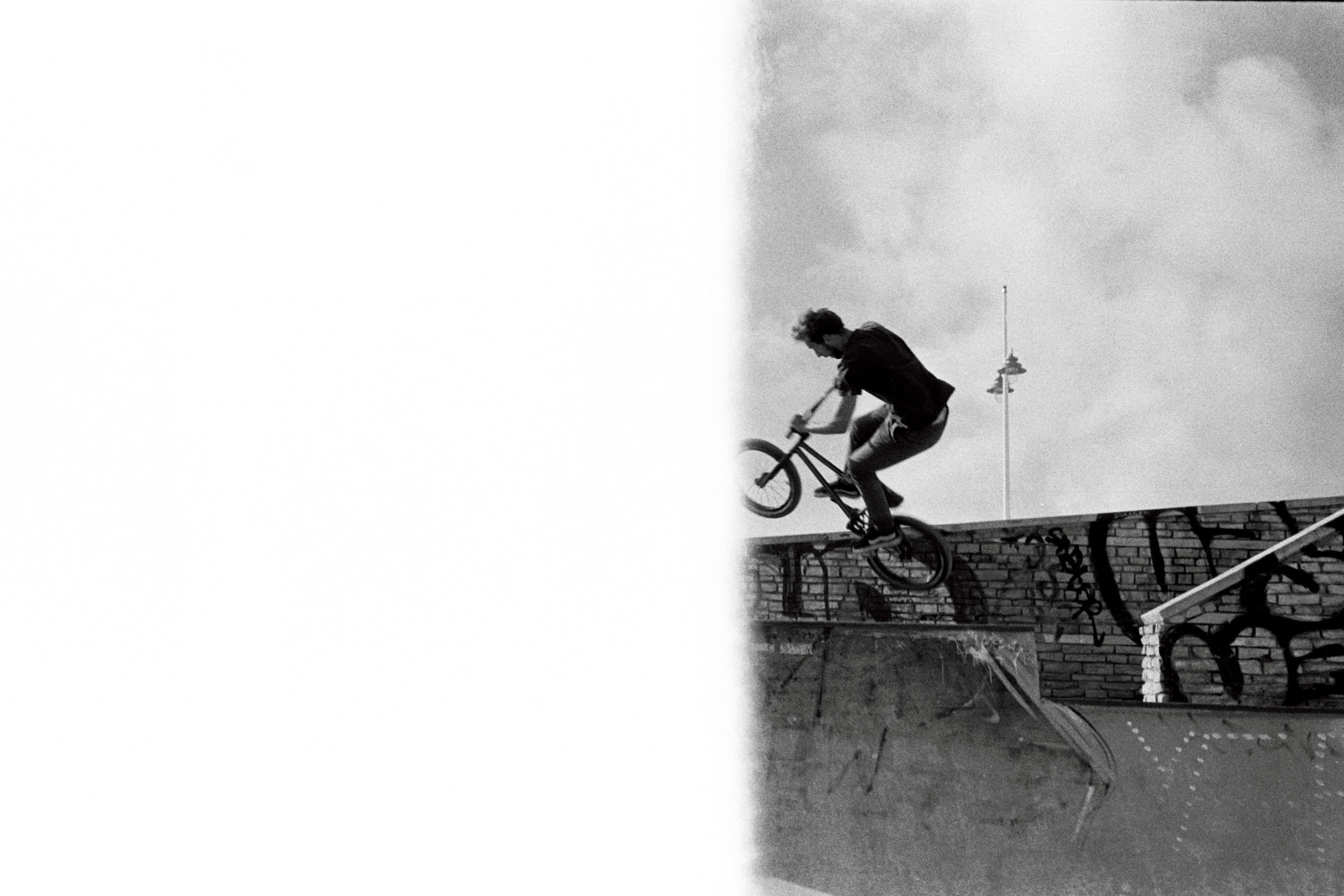 un rider en BMX effetue un saut dans un skatepark , on distingue une bande blanche sur la partie gauche de l'image argentique
