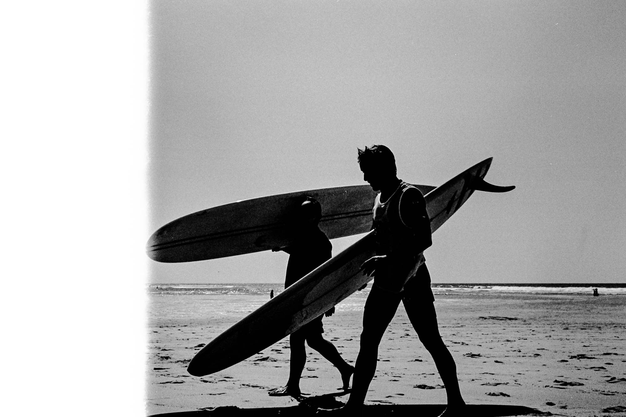 deux surfeurs en noir et blanc marchent sur la plage et portent leurs planches en bord d'océan. une photographie argentique avec une bande visible en partie gauche
