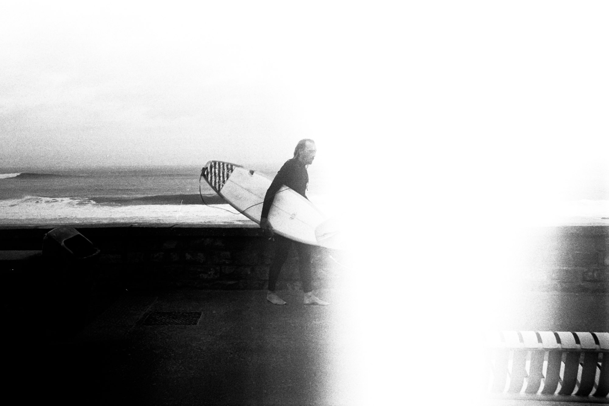 un surfeur marche piued nu dans la rue devant le port de capbreton. une bande blanche due à une erreur de pellicule est visible en son milieu