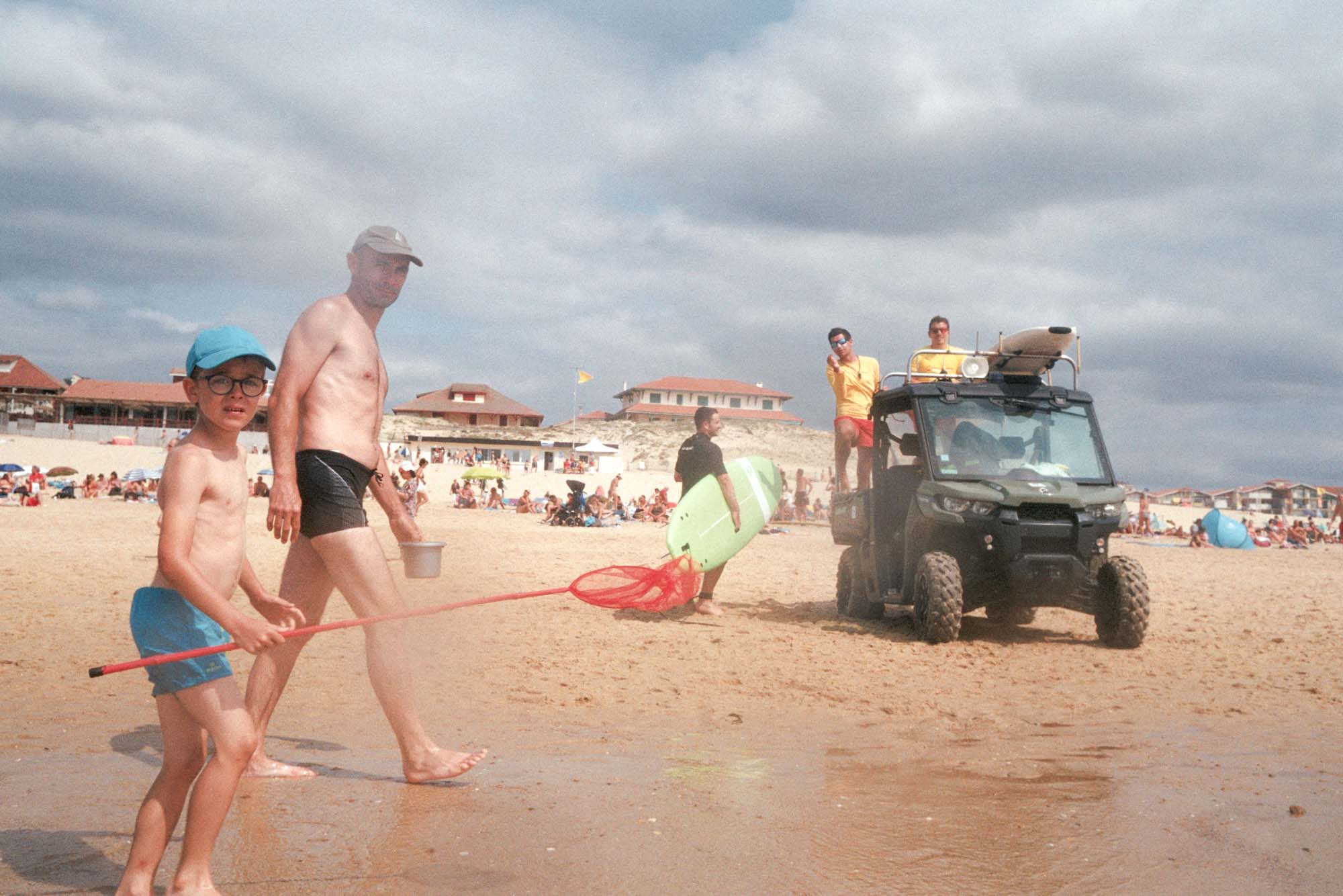 un père et son fils marchent torse nu avec une épuisette et me regardent les prendre en photo argentique, on peut voir les MNS derrière en t-shirt jaune des sauveteurs.