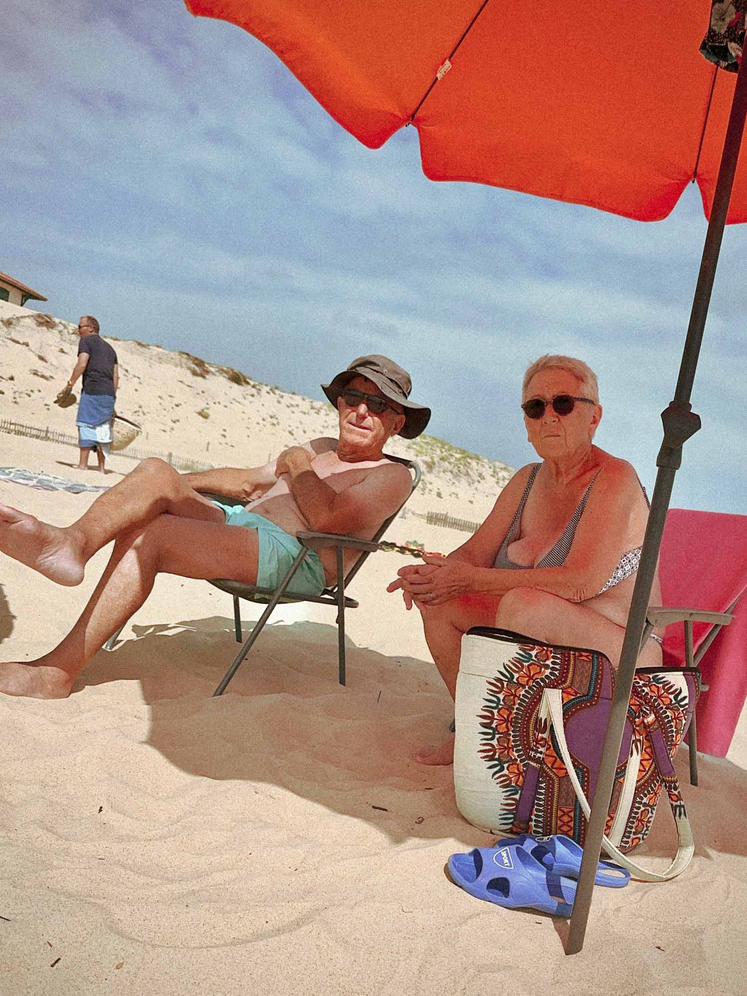 un couple agé me regardent les prendre en photo argentique au LEICA M6. Ils sont sous un parasol rouge, assis sur des chaises de camping et l'homme porte jun chapeau