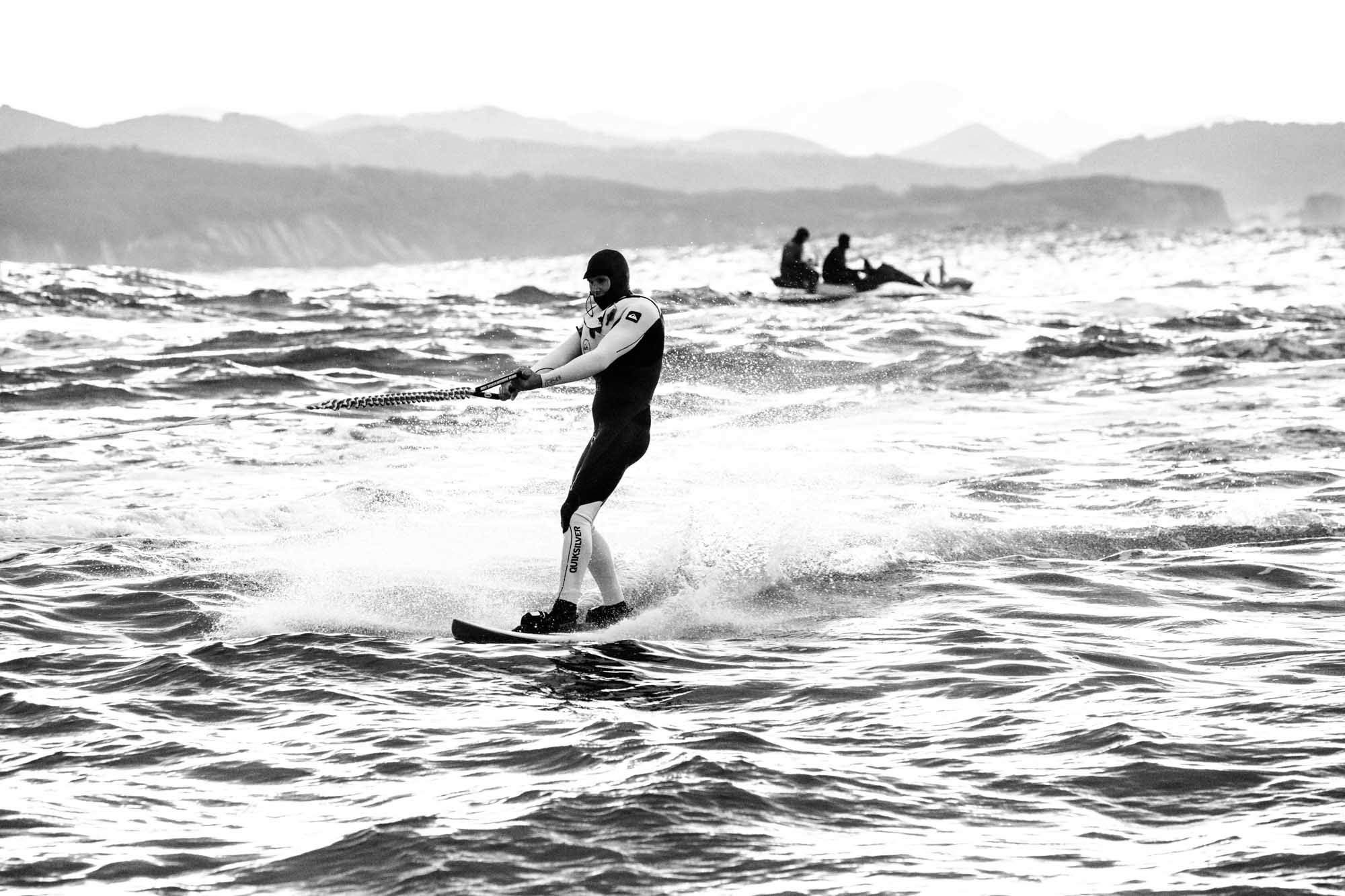 un surfeur tracté prend son élan pour surfer belharra, on distingue la montagne en fond et un jet ski, photographie noir et blanc