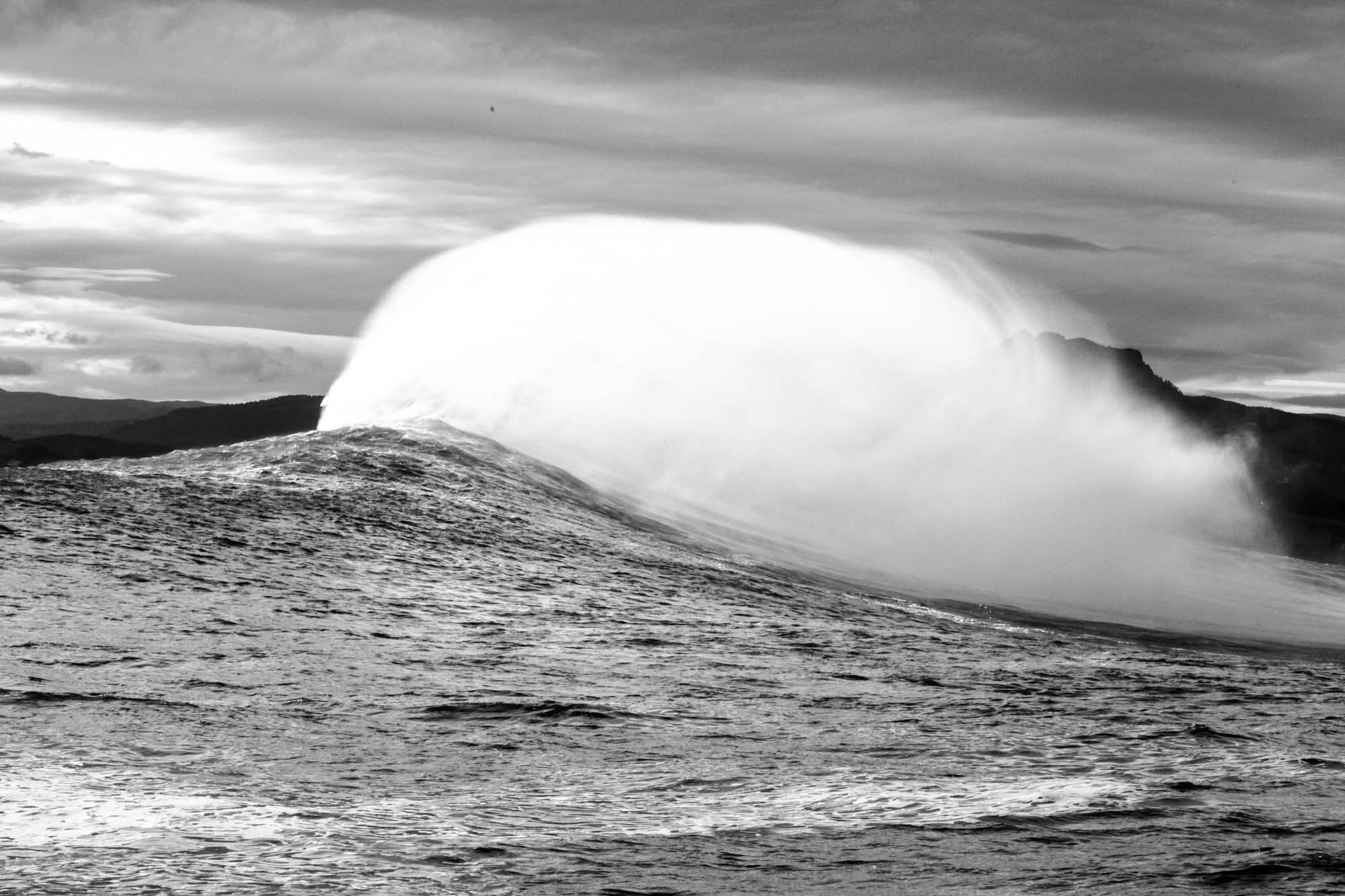 vue de profil de la vague de belharra avec un gros spray en l'air et les montagnes basques derrière en noir et blanc
