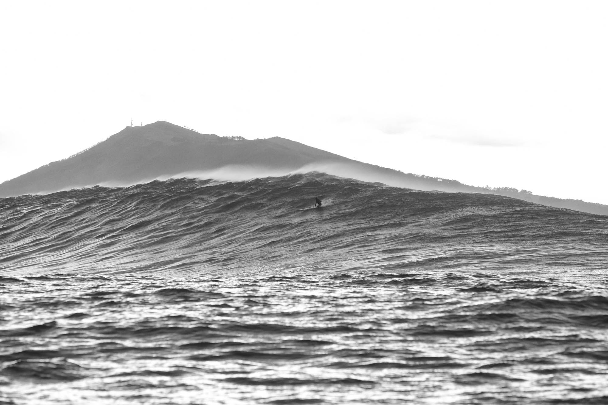 une vague et la montagne basque en noir et blanc, un surfeur en haut assis sur sa planche attend la bonne