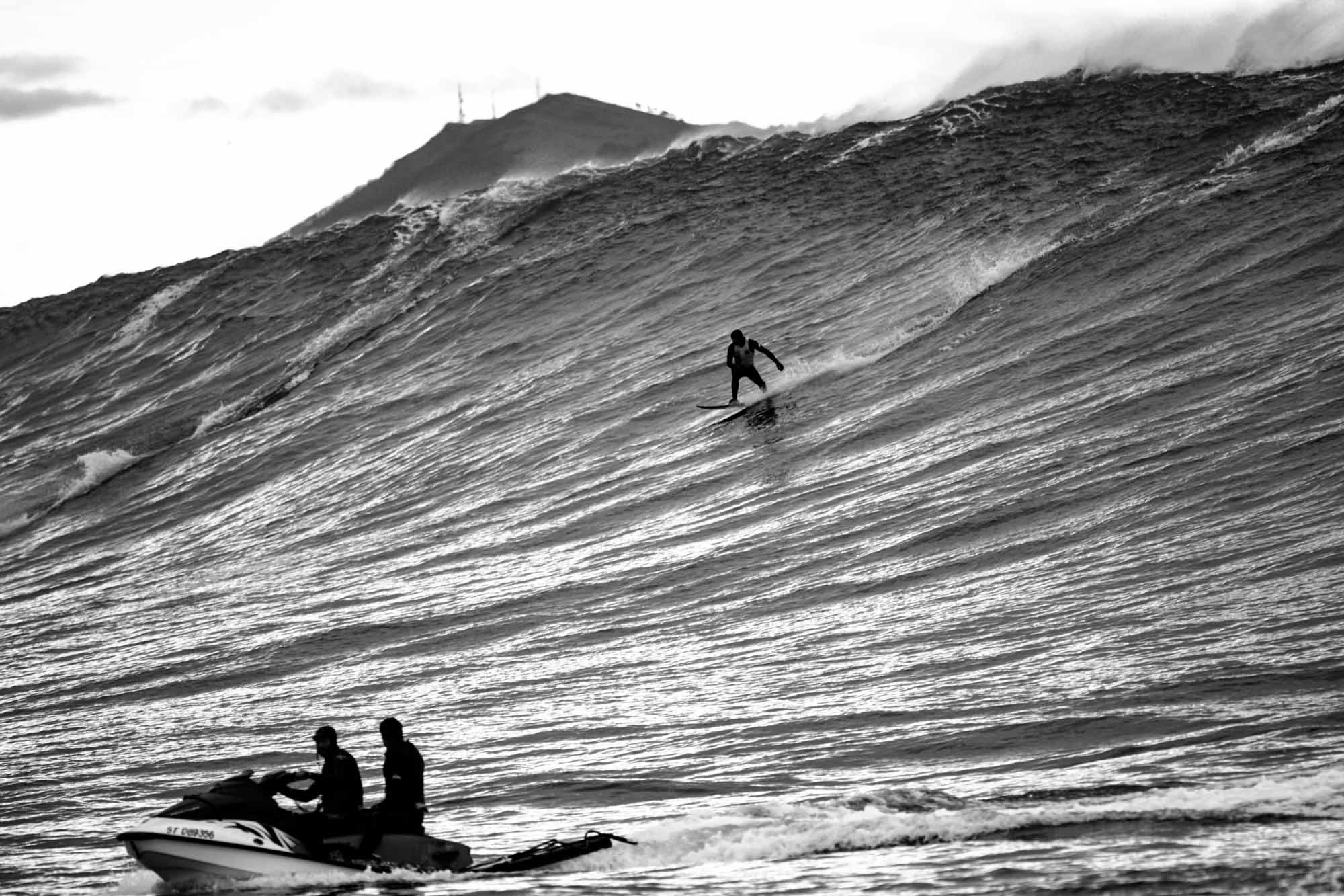 un surfeur glisse sur la pente de la vague géante et son jegt ski le suit en gros plan, on distingue encore la montagne basque, la rune
