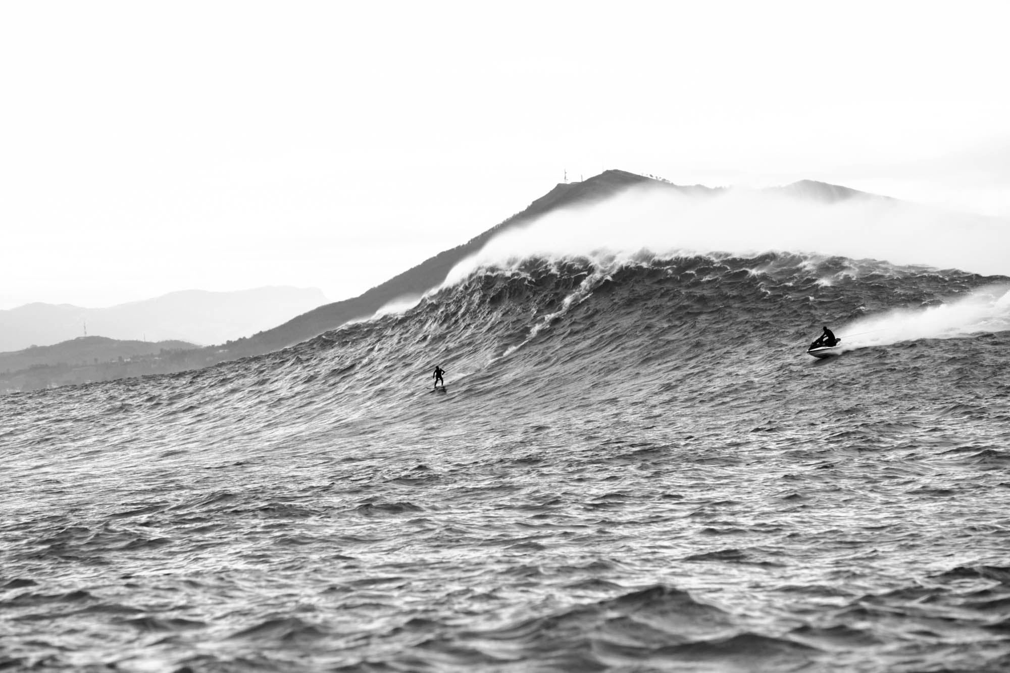 un surfeur descend la vague géante de belharra, photographie de profile en noir et blanc avec la rune derrière et un jetski qui suit le surfeur par sécurité