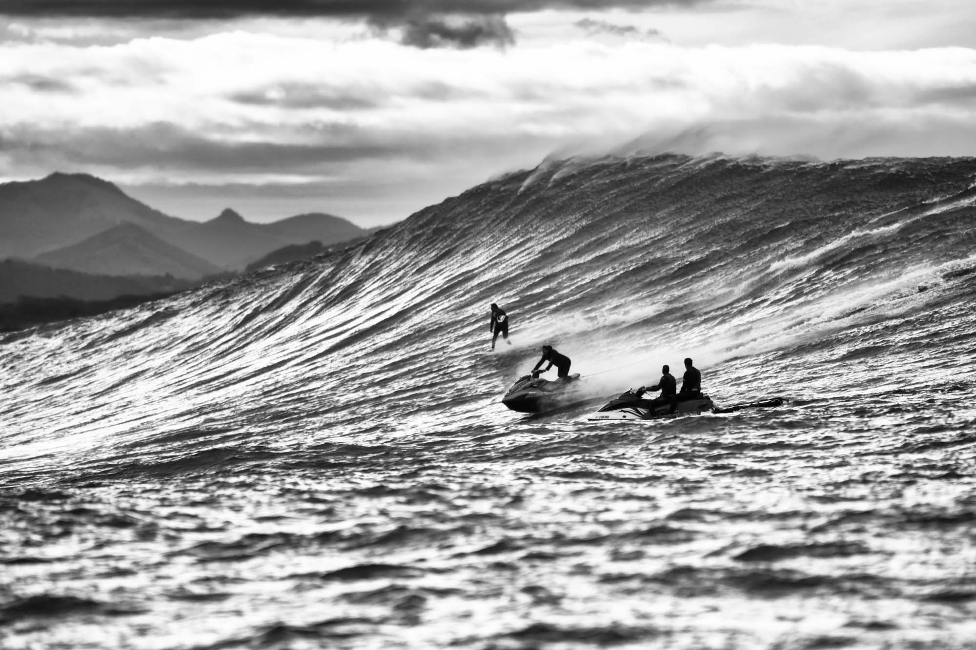 un surfeur descend la vague de belharra de 15 mètres, il est de profil et suivi par son jet ski prêt à àintervenir, la sensation de vitesse est là