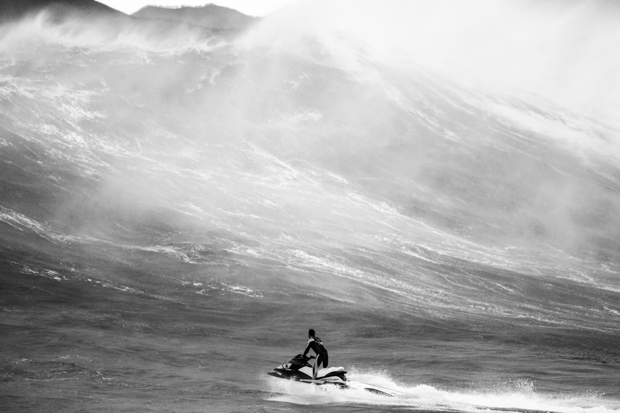 on voit un mur d'eau énorme avec un spray représentant l'arriére de la vague de 15 mètres et un jet ski qui a laché son binome et se prépare à aller le récupérer