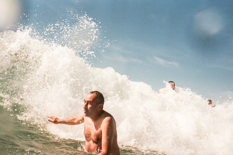 un homme essaie d'esquiver une vague qui arrive sur lui mais qui semble trop grosse, la mousse derière esdt assez énorme et il ne regartde pas la vague, photographie couleur argentique