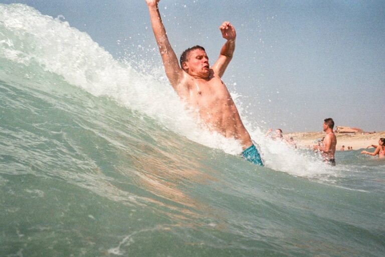 photographie couleur argentique d'un jeune adulte qui glisse sur une vague debout avce le corps tordu, brasd en l'air au bord des vagues