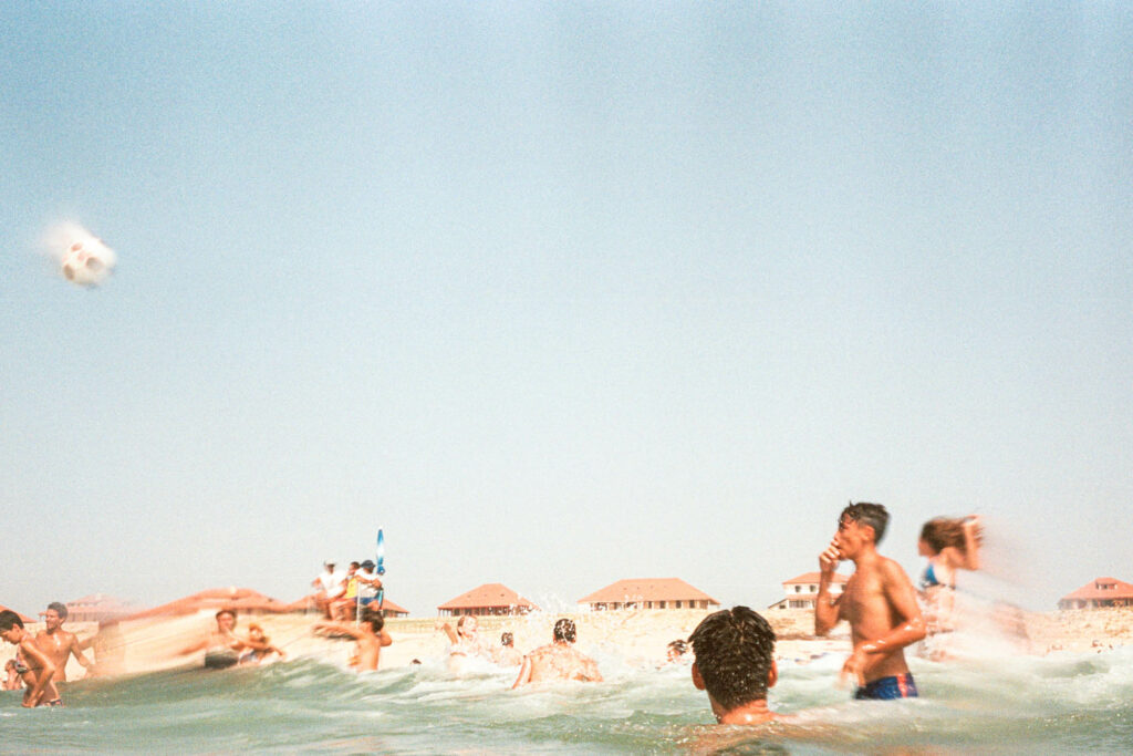 Scène floue de jeunes jouant au ballon dans l'eau face aux maisons de bord de mer, tirage d'art argentique couleur "Foot de Plage", MonVilain