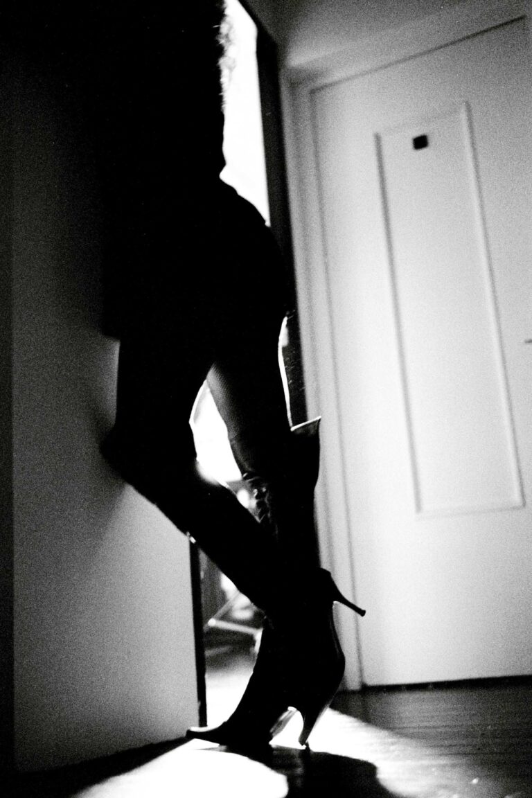 une silhouette féminine en botte à talon s'appuie contre un mur